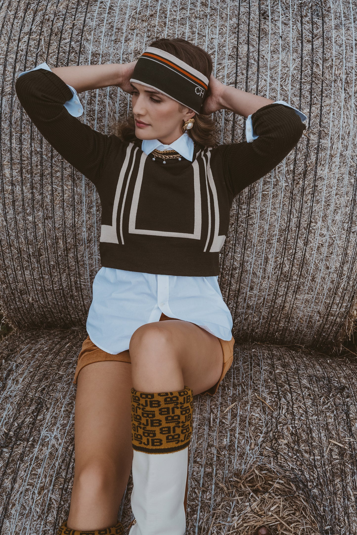 A woman posing in a straw-colored straw hat with a striped band, a dark brown knit sweater with cream geometric stripes, a light blue collared shirt, a brown mini skirt, and white knee-high socks with a brown pattern, seated against a straw hay bale background.
