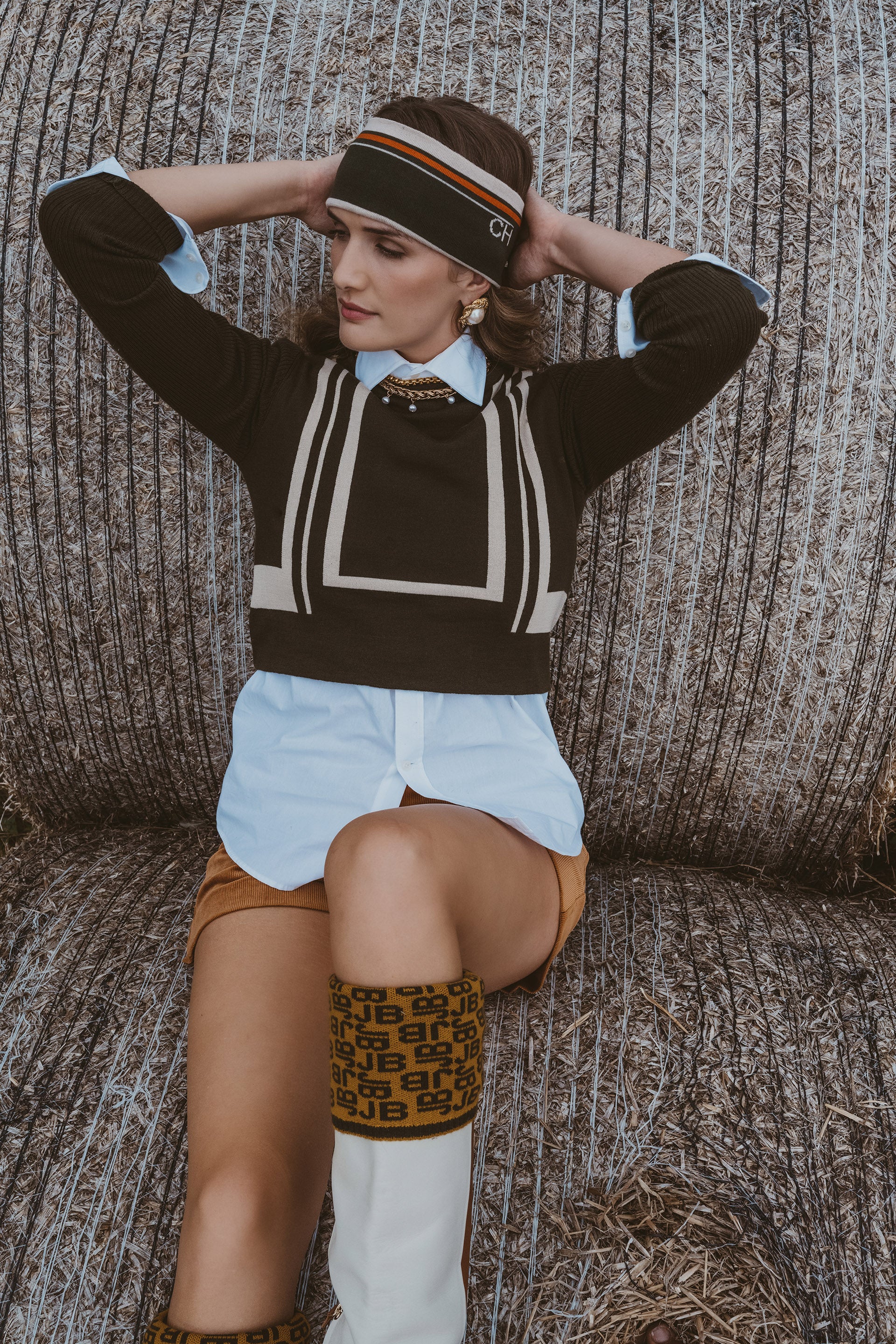 A woman posing in a straw-colored straw hat with a striped band, a dark brown knit sweater with cream geometric stripes, a light blue collared shirt, a brown mini skirt, and white knee-high socks with a brown pattern, seated against a straw hay bale background.